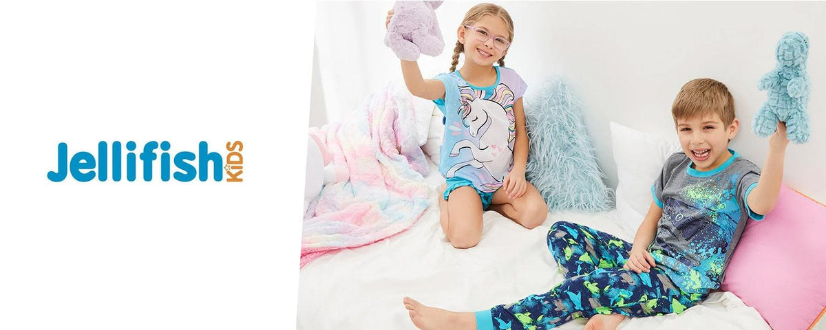 Two kids in colorful pajamas sitting on a bed holding plush toys, cozy bedroom setting