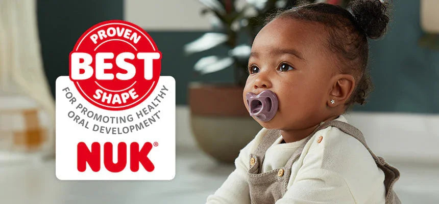 Baby girl with a purple pacifier sitting indoors beside a NUK pacifier advertisement sign.