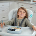 Infant high chair with smiling toddler using fork, eating fruit and yogurt in a modern kitchen.
