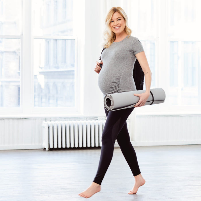 Maternity activewear leggings and t-shirt worn by a pregnant woman holding a yoga mat indoors.
