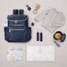 Navy diaper bag with baby essentials: striped cardigan, pacifier, bottle, teether, diaper, changing mat, and food container