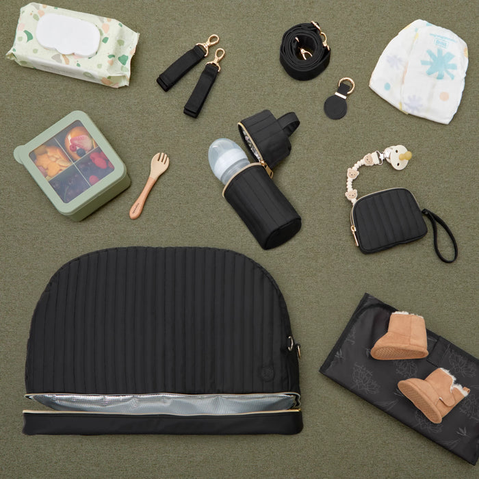 Flat lay of baby essentials including black diaper bag, bottle holder, pacifier clip, wipes, diaper, bento box, baby boots and feeding fork on green surface.