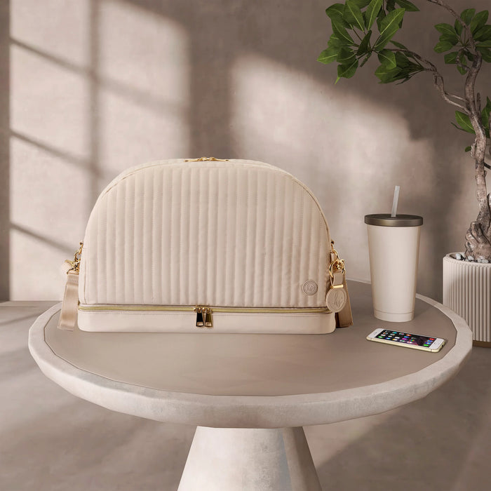 Beige quilted diaper bag with gold zippers on table, next to tumbler and smartphone.
