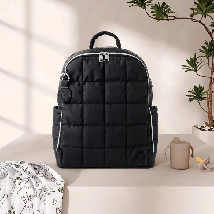 Black quilted diaper backpack with baby bottle, pacifier, and swaddle on beige table