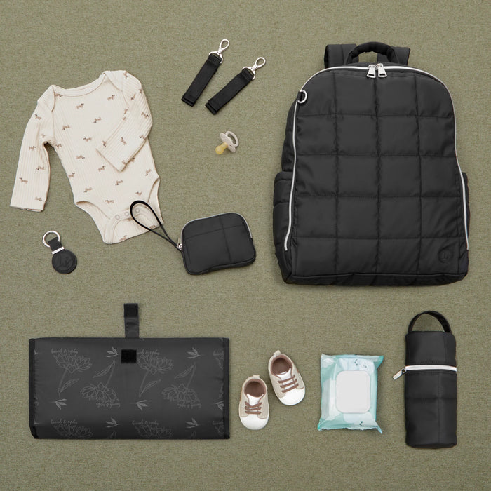 Baby diaper bag essentials set with black backpack, changing mat, infant onesie, pacifier, baby shoes, wipes, and accessories on green background