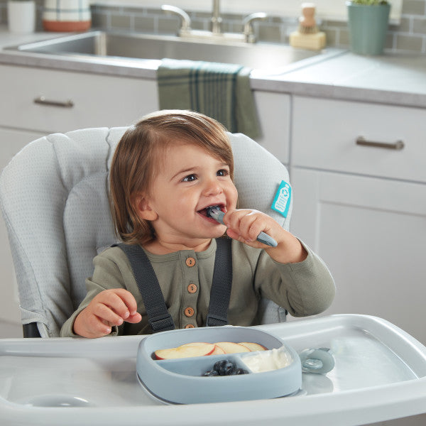 Baby self-feeding with silicone spoon in high chair, using divided suction plate in kitchen setting.