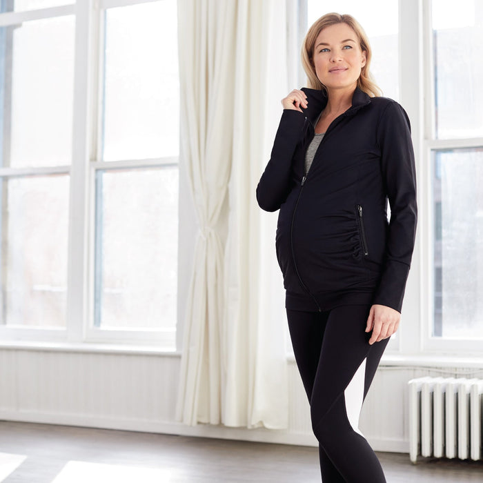 Maternity activewear jacket and leggings worn by a pregnant woman standing in a bright room.