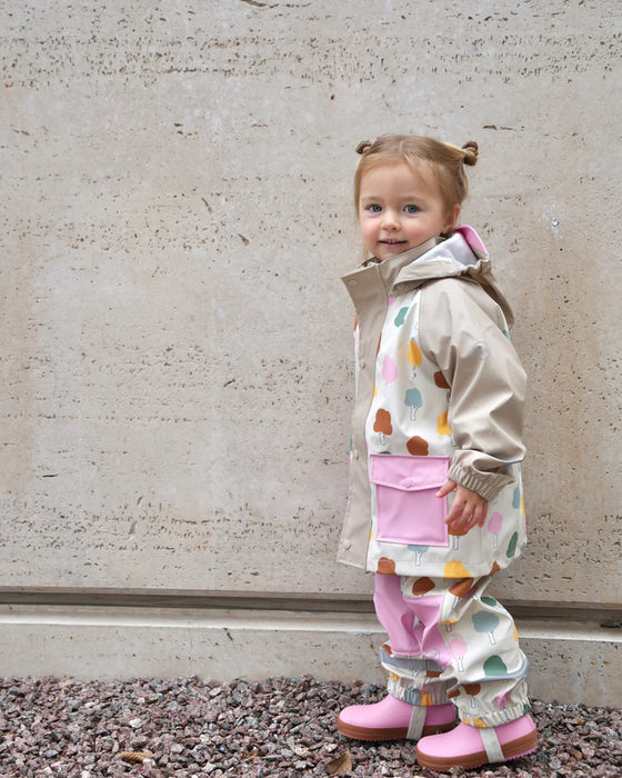 Toddler in a beige rain jacket with colorful tree print and pink boots standing outdoors on gravel.