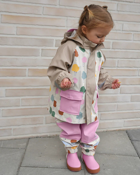 7AM rain jacket with colorful tree print, worn by a young child standing outdoors in pink boots.