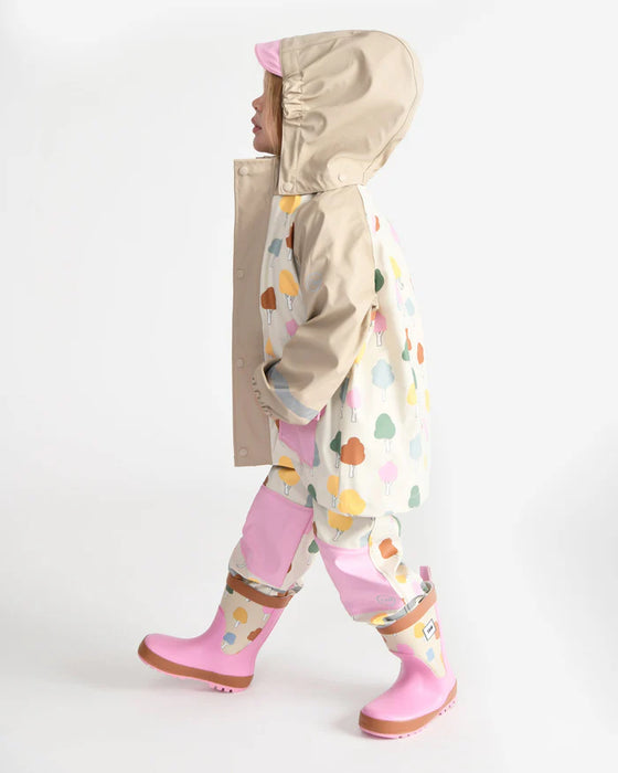 Child wearing a beige hooded rain jacket with colorful tree pattern, matching pants and pink boots.