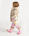 Child wearing a colorful rain jacket, patterned pants, and pink rain boots walking indoors.