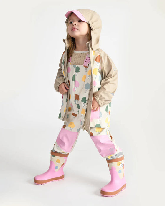 Child wearing a beige hooded rain jacket, patterned overalls, and pink rain boots standing indoors.