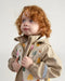 7AM rain jacket in beige with colorful tree print, worn by a young child facing upward indoors.