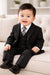 Smiling baby in black formal suit, white shirt, and striped tie sitting on a sofa