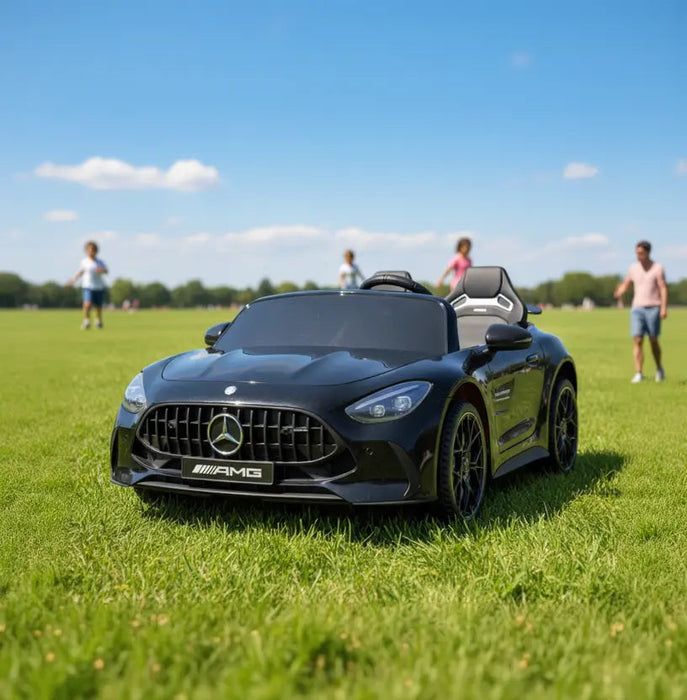 AMG kids ride-on electric car in black shown on grass in a park with children playing nearby.