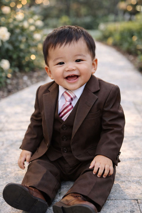 Baby brown suit set with tie, smiling infant sitting on a garden pathway outdoors.