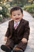 Baby brown suit set with tie, smiling infant sitting on a garden pathway outdoors.
