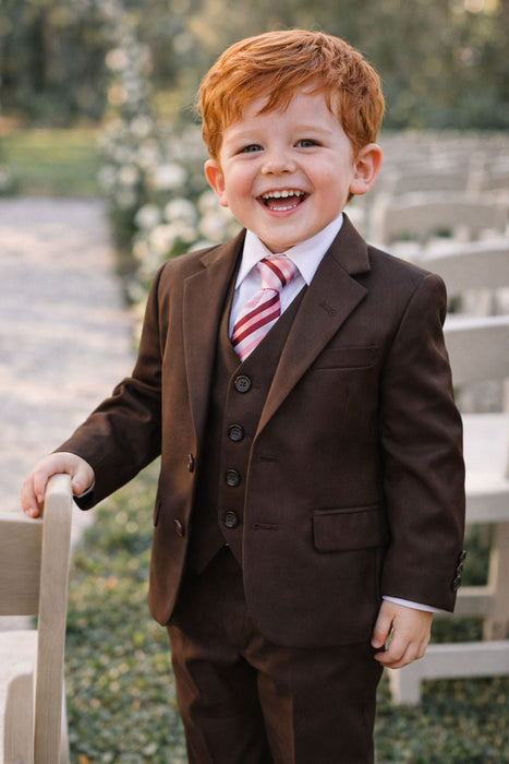 Toddler brown suit with vest and striped tie, smiling outdoors beside chairs at an event.