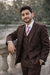 Brown suit for young men shown worn outdoors against a stone railing, paired with striped tie.
