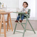 Toddler sitting in green high chair with tray, eating at dining table in modern kitchen