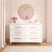 White dresser with gold handles, round mirror, pink flowers, and decorative vases in a nursery room