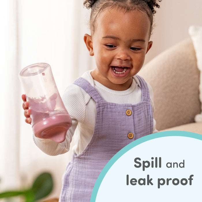 Toddler holding a spill-proof sippy cup, smiling in a cozy living room setting.