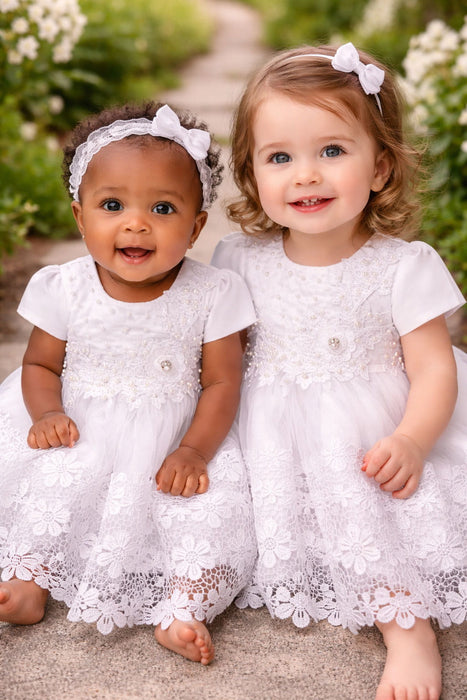 White lace baby dresses shown on two young girls sitting outdoors on a garden path.