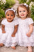 White lace baby dresses shown on two young girls sitting outdoors on a garden path.