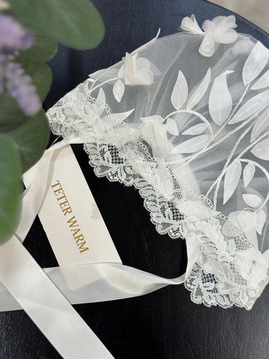 Teter Warm baby lace bonnet with 3D leaf details and ribbon ties displayed on a dark surface.