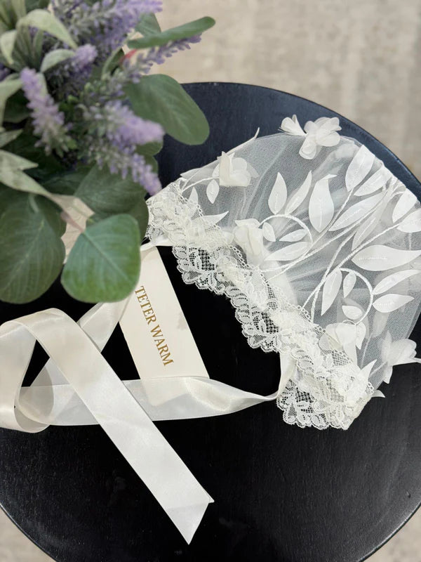 Teter Warm AB06 bonnet with 3D leaves and lace trim displayed on a black table beside flowers.