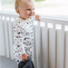 A smiling toddler stands by a white railing wearing a white long-sleeve pajama sleeper with car print.