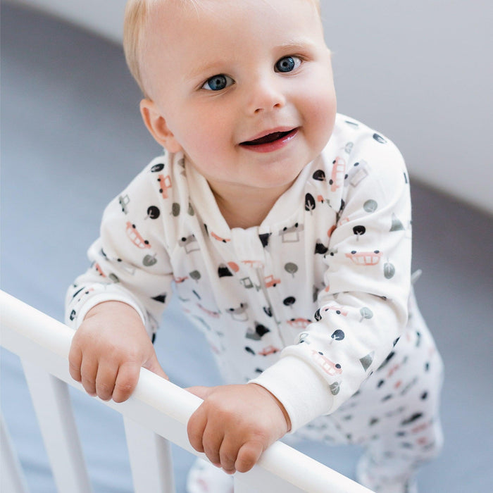 A smiling baby wearing a white long-sleeve pajama with car pattern, leaning on a white crib rail.