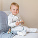 A baby sitting on a sofa wearing a white long-sleeve footed sleeper with a gray animal print, holding a wooden toy