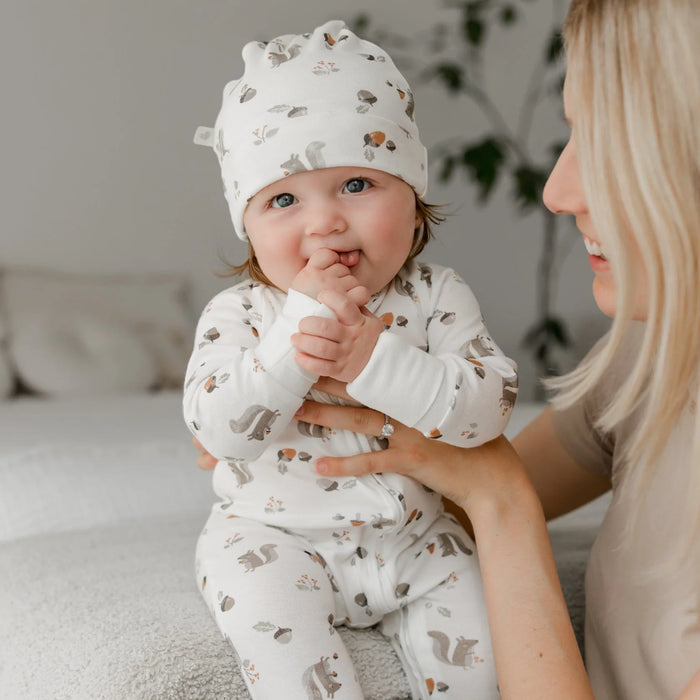 Baby wearing white bamboo sleeper pajamas and hat with squirrel print, held by smiling woman
