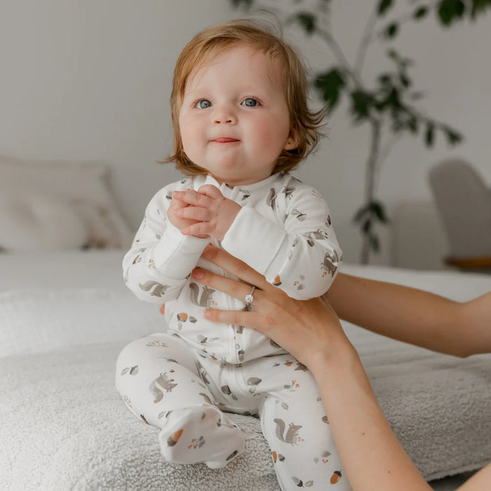 Baby in white bamboo sleeper pajamas with squirrel print sitting on bed indoors
