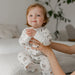 Baby in white bamboo sleeper pajamas with squirrel print sitting on bed indoors