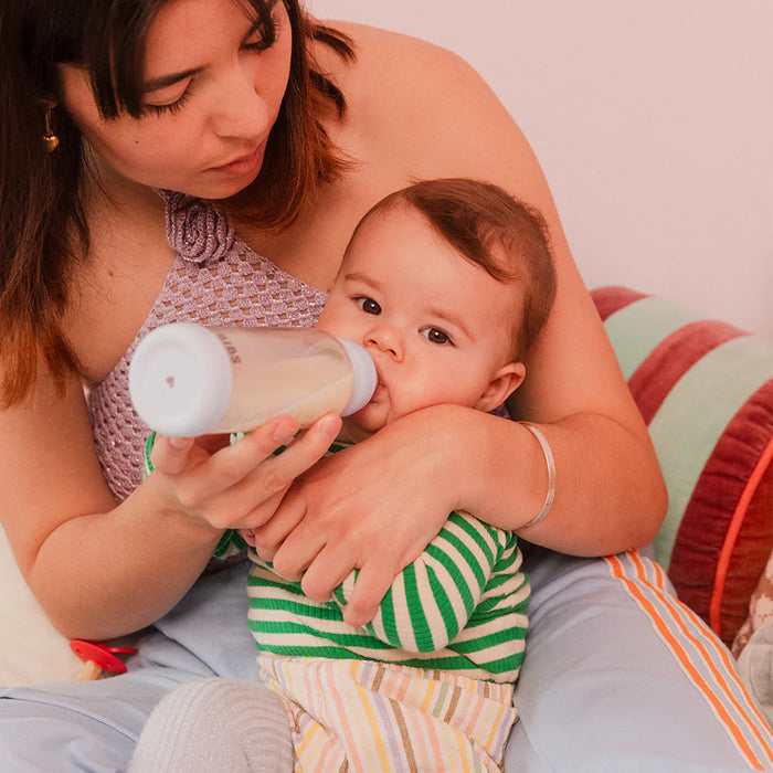 Baby glass bottle being used by a woman to feed an infant sitting on her lap at home.