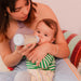 Baby glass bottle being used by a woman to feed an infant sitting on her lap at home.