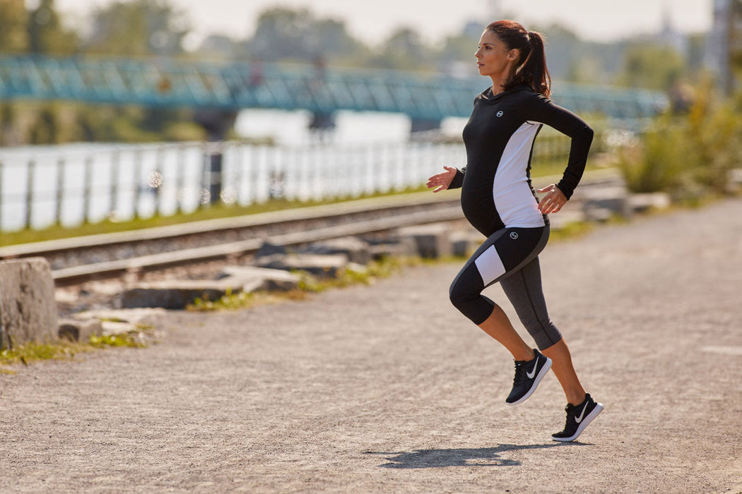 Maternity activewear set worn by a pregnant woman jogging outdoors on a sunny path near railroad tracks.