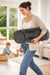 BabyBjörn bouncer seat folded and carried by a woman in a bright living room with a child playing nearby.