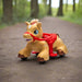 Children’s ride-on horse toy with red saddle and wheels shown outdoors on a forest path.