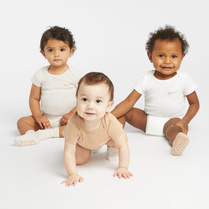 Nike baby onesies in beige and white shown on three babies sitting and crawling on a white background.
