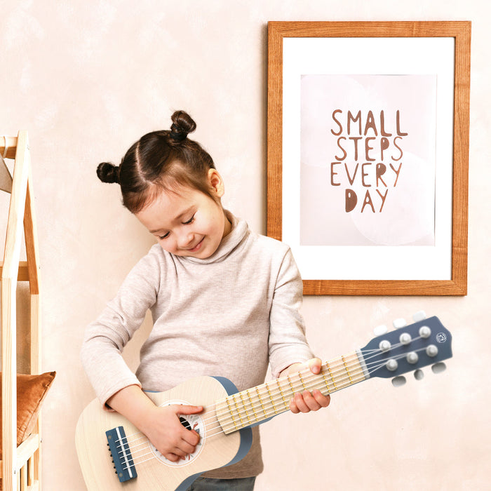 Child-sized wooden guitar being played by a toddler in a cozy room with wall art in the background.