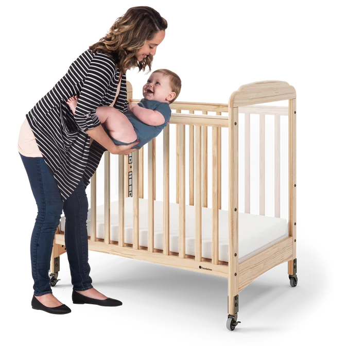Woman placing smiling baby in natural wood crib with clear side panel and wheels