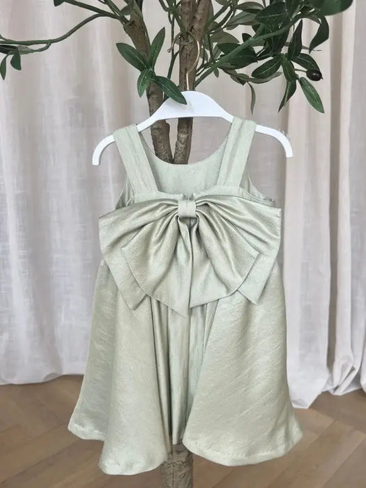 TETER WARM Clarice Sage Green Party Dress with large bow on hanger against neutral curtains.