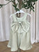 TETER WARM Clarice Sage Green Party Dress with large bow on hanger against neutral curtains.