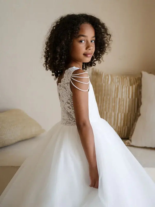 Teter Warm Lorelai Pearl Communion Dress shown on a young girl in a softly lit room with cream pillows.