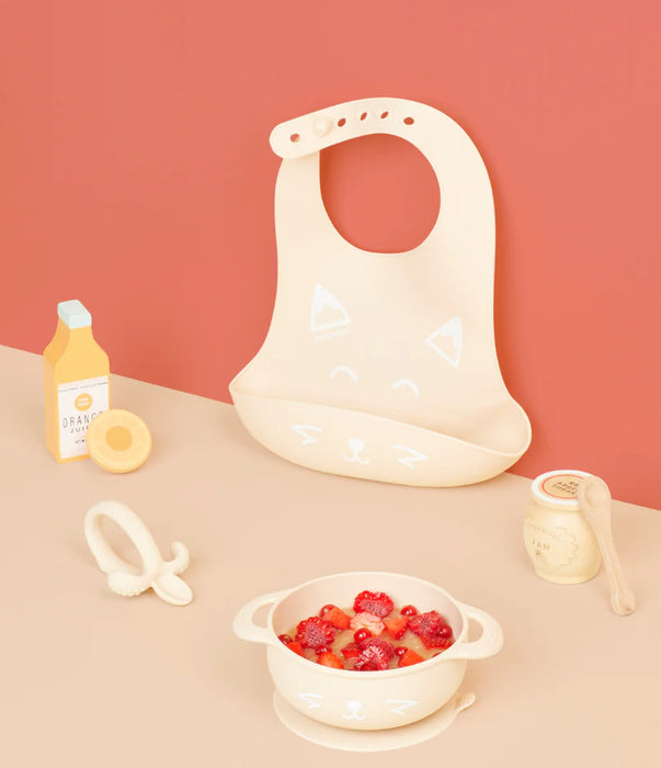 Cute animal face silicone baby bib, bowl with strawberries, baby spoon, teether, and play juice bottle on peach background.