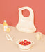 Cute animal face silicone baby bib, bowl with strawberries, baby spoon, teether, and play juice bottle on peach background.