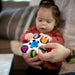 b.box baby sensory toy being held by an adult as a baby explores it in a living room setting.